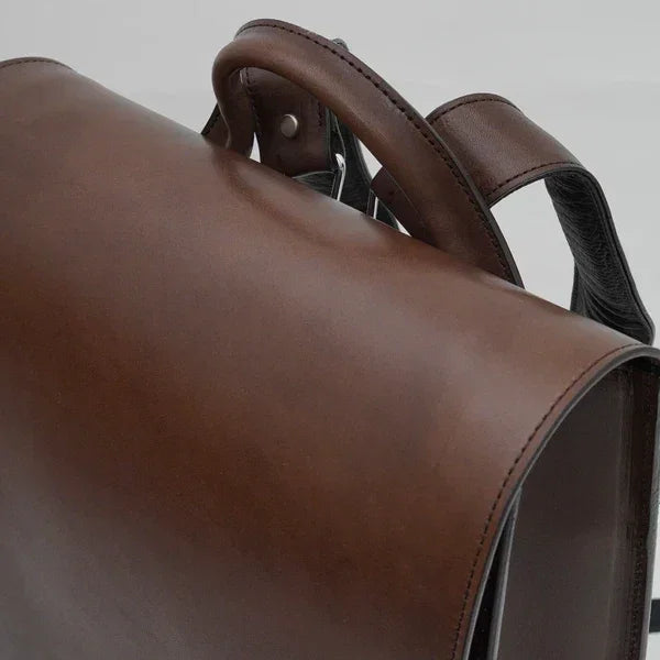 Close-up of brown leather briefcase handle and flap with detailed stitching against a plain background