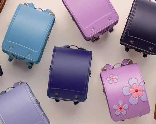 Colorful Japanese school backpacks arranged on a light surface, featuring solid and floral designs in blue, purple, and lavender