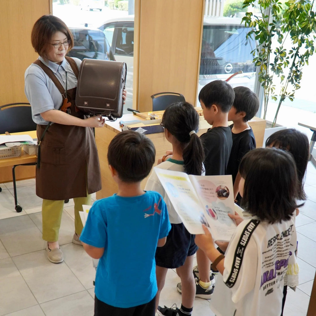 Teacher showing a brown leather backpack to a group of children holding brochures in a bright classroom